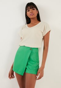 Haut beige texturé à manches courtes associé à un short taille haute vert vif avec une fermeture à bouton sur le côté et un ourlet asymétrique.