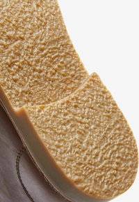 Suela de goma con una superficie texturizada en color beige, con forma redondeada y una ligera curvatura, unida a una parte superior de cuero con costuras visibles.