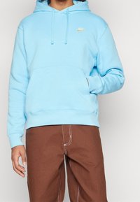 Persona vistiendo un sudadera de Nike azul claro con bolsillo frontal y pantalones marrones con costuras blancas visibles, con la mano parcialmente en el bolsillo.