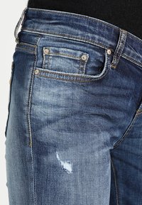 Vaqueros de denim azul oscuro con un acabado desteñido y desgastado, que presentan un pequeño rasguño, diseño de cinco bolsillos y costuras en contraste.