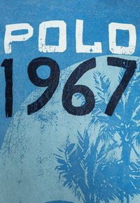 T-shirt in cotone blu caratterizzata dal testo "POLO 1967" in bianco e blu scuro, con una grafica di una palma e dettagli in superficie testurizzati.