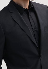 Uomo che indossa una giacca nera sopra una camicia nera, abbottonata senza cravatta, su uno sfondo chiaro.