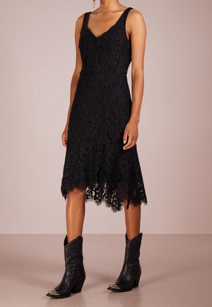 Femme portant une robe midi en dentelle noire sans manches avec un ourlet festonné, associée à des bottes de cowboy en cuir noir avec des bouts métalliques.
