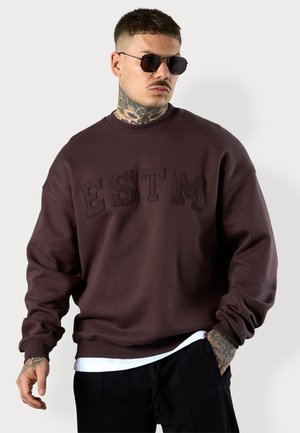 Dark brown sweatshirt with "ESTM" embossed in darker thread, round neckline, dropped shoulders, and ribbed cuffs. Worn over a white shirt.