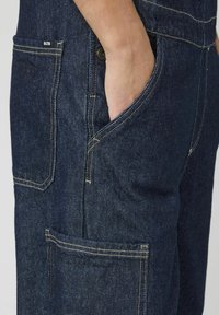 Salopette en denim foncé avec une poche latérale, des coutures jaunes contrastantes et une coupe décontractée. Le matériau présente une finition tissée texturée.