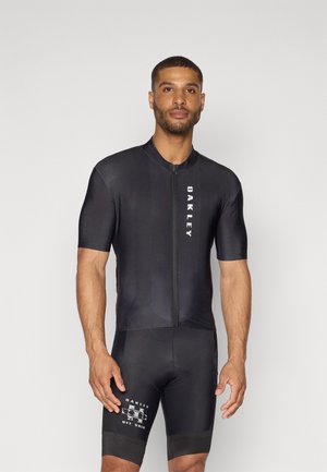 Ensemble de cyclisme noir avec des manches courtes, fermeture éclair sur le devant et branding Oakley. Le tissu est lisse et ajusté, avec des détails en panneaux sur les shorts.