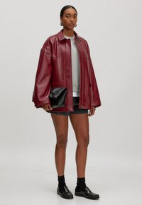 Rode leren oversized jas met een puntige kraag, twee voorzakken en een ritsluiting, gecombineerd met een grijs sweatshirt en zwarte accessoires.