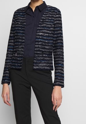 Femme portant un blazer texturé à rayures noires, blanches et bleues par-dessus une chemise boutonnée bleu foncé et un pantalon noir, debout devant un fond uni.