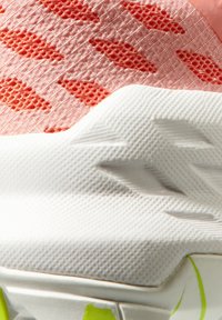 Gros plan sur la semelle intermédiaire blanche texturée d'une basket et sur la tige en mesh rose avec des trous d'aération hexagonaux orange.