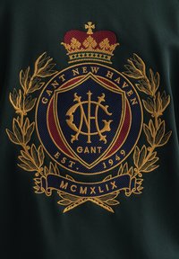 Tela verde con un escudo bordado en tonos dorado y burdeos. El diseño incluye una corona, hojas de laurel y elementos de texto.
