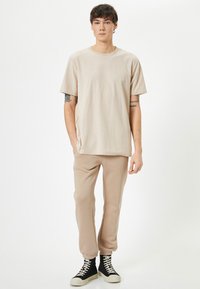 Ung mand står iført en beige kortærmet skjorte, matchende joggerbukser, sorte højhælede sneakers og viser synlige tatoveringer på begge arme.