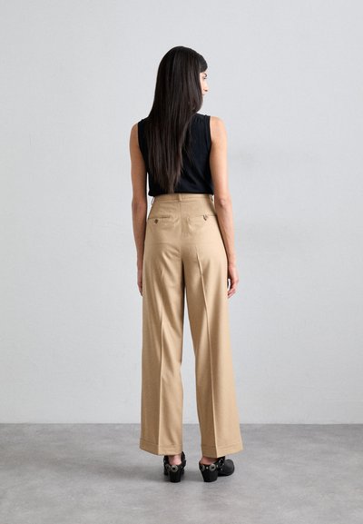 Helle weite Hose mit hoher Taille, ausgestattet mit zwei Gesäßtaschen und flachem Vorderdesign. Kombiniert mit einem schwarzen ärmellosen Oberteil und hohen Schuhen.