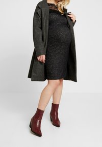 Abito da maternità nero fino al ginocchio con sovrapposizione in pizzo, abbinato a un cappotto grigio scuro. Stivali alla caviglia bordeaux con finitura testurizzata e tacco a blocco.