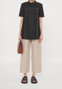 Persona con una camicia nera a maniche corte con bottoni, pantaloni beige a gamba larga, sandali neri e una piccola borsa marrone.