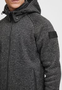 Dark gray zip-up hoodie with a textured knit fabric, adjustable hood, and side pockets. Features a black label on the shoulder.