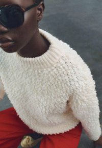 Weißer strukturierten Pullover mit geripptem Kragen, kombiniert mit roten Hosen. Das Modell trägt dunkle Sonnenbrillen, wodurch eine Nahaufnahme des Outfits zu sehen ist.