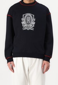 Sudadera de la Marina con acentos rojos y emblema blanco que presenta el texto "Hilfiger". Puños y cuello acanalados, tela suave, ajuste relajado.