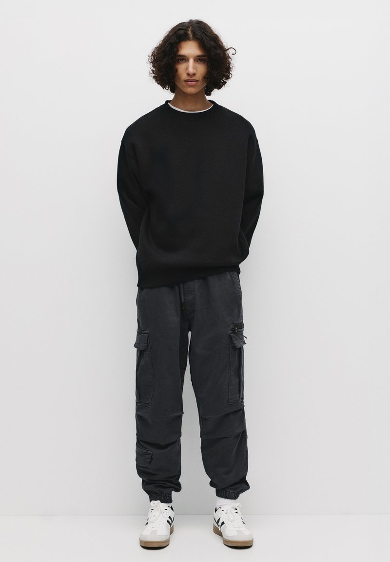 Hanorac oversized negru, pantaloni cargo închis cu buzunare laterale și încălțăminte albă cu accente maro. Textură netedă, design casual.