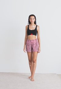 Čierny crop top s červeným akcentom, skombinovaný s ružovými šortkami s leopardím vzorom. Modelka stojí bosá proti neutrálnemu pozadiu.