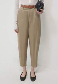 Pantalons beiges taille haute, avec des plis à l'avant et une ceinture marron. Associés avec un haut blanc ajusté à côtes et des chaussures plates noires.