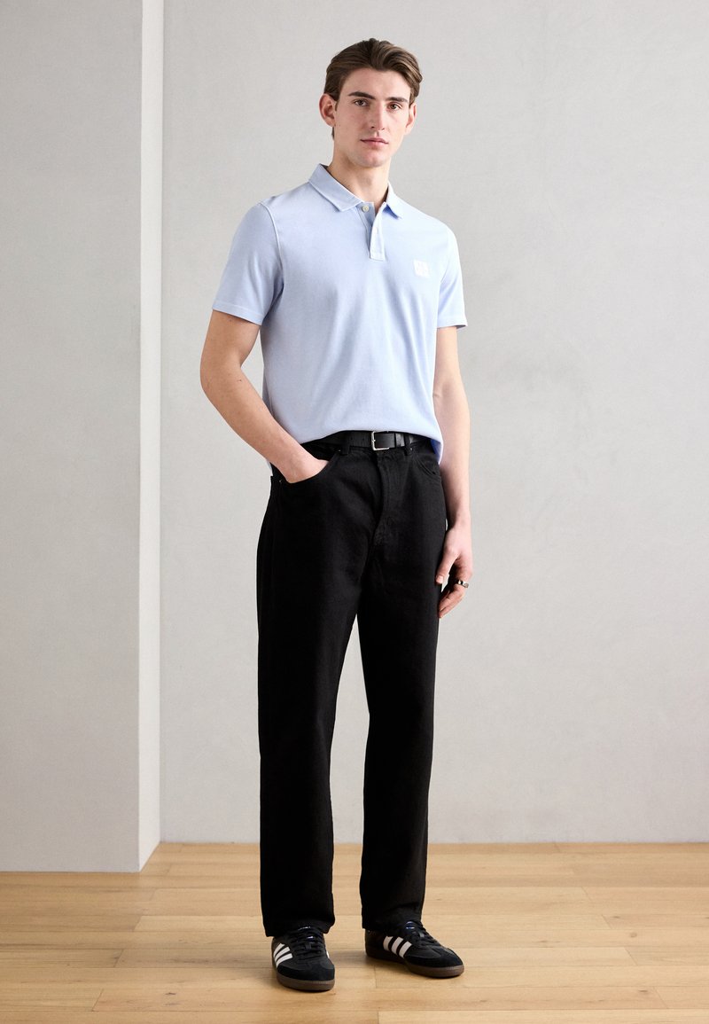 Helles blaues Polohemd mit kurzen Ärmeln, mit Kragen, getragen mit schwarzen Hosen und schwarzen Sneakers mit weißen Streifen.