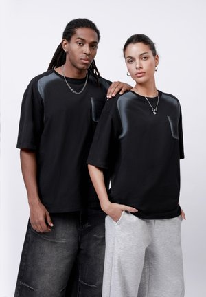 Schwarze übergroße T-Shirts mit satinierten Schulterakzenten und Fronttaschen, kombiniert mit hellgrauen, weit geschnittenen Sweatpants.
