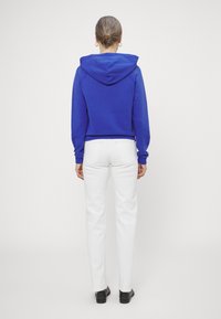 Zugeschnittenes blaues Hoodie mit Kapuze, langärmlig, getragen mit weißen geraden Jeans und schwarzen Schuhen. Schlichter Hintergrund.