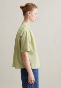 Camicetta verde chiaro a maniche corte con colletto rialzato, vestibilità comoda e pieghe sottili. Abbinata a una gonna di jeans blu scuro.