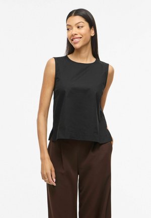 Kvinne iført svart ermeløs topp og brune bukser, smilende og ser litt mot venstre mot en ensfarget hvit bakgrunn.