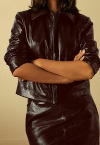 Veste en cuir bordeaux avec une fermeture éclair, un large col et un design court. Texture brillante. Associée à une jupe en cuir assortie.