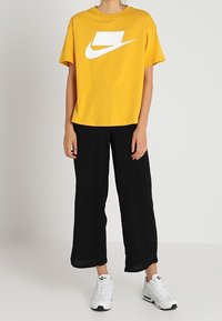 Keltainen puuvillapaita, jossa on valkoinen Nike-logo, yhdistettynä löyhiin mustiin housuihin ja valkoisiin lenkkareihin, esittelee rentoa urheiluvaatemuotoilua.