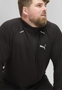 Schwarzes langärmliges Sportoberteil mit halbem Reißverschluss am Kragen, mit weißen Akzenten und einem kleinen weißen Puma-Logo auf der Brust. Glatte Stofftextur.