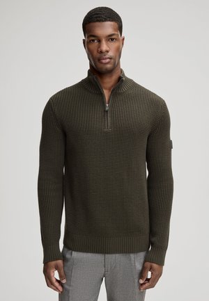 Mann trägt einen dunkelolivgrünen, strukturierten Strickpullover mit Viertel-Reißverschluss-Kragen und graue Hose, steht vor einem schlichten hellen Hintergrund.