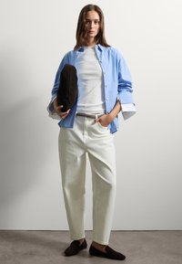 Donna con lunghi capelli castani che indossa una camicia a righe azzurre chiara sopra una maglietta bianca, pantaloni bianchi, mocassini scuri, mentre tiene in mano una borsa a pochette nera.