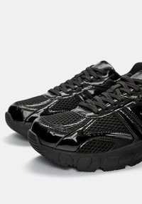 Black athletic shoes with a mix of glossy patent leather and textured mesh. Features prominent laces and a rugged rubber sole.