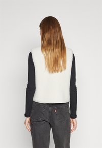 Faux fur-väst i off-white med ärmlös design, i kombination med en svart långärmad topp och mörkgrå jeans med bakfickor.