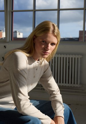 Jeune femme aux cheveux blonds portant un pull beige à boutons et un jean bleu, assise à l'intérieur près d'une grande fenêtre avec des bâtiments de la ville à l'extérieur.