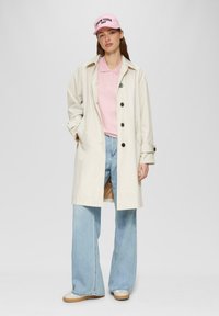 Beige trenchcoat met zwarte knopen, roze polo, lichtblauwe wijde jeans en een roze pet met de tekst "NEW YORK 1987".