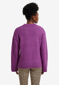 Frau mit kurzen lockigen Haaren, die einen lila Strickpullover und beige Leopardenmuster-Hosen trägt, von hinten gegen einen weißen Hintergrund gerichtet.
