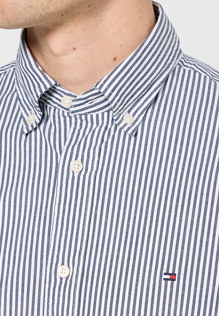 Chemise boutonnée à rayures verticales bleu et blanc, avec un col, des boutons blancs et un petit logo sur la poitrine. Texture en tissu de coton.