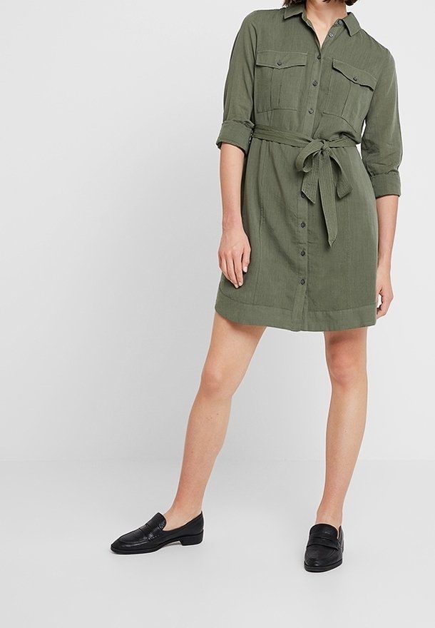 Khaki šaty košilového střihu s knoflíkovým zapínáním, dvěma náprsními kapsami, odnímatelným páskem v pase a ohrnutými rukávy. Nosené s černými mokasíny.