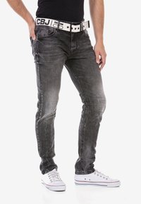 Slim-fit zwarte denim jeans met een vervaagde textuur, gecombineerd met een witte riem met zwarte letters en witte sneakers.