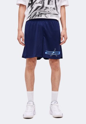 Personne portant un short de basketball bleu marine avec des rayures bleu clair et un logo Jumpman, des chaussettes blanches, des baskets blanches et un t-shirt graphique blanc.