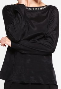 Haut noir à manches longues en tissu brillant avec un col rond. Détails remarquables : des ornements le long du col. Design simple et décontracté.