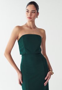 Zaļa strapless kleita, izgatavota no pieguļoša auduma, ar taisnu dekoltē un plūstošu siluetu, apvienota ar zelta rotām.