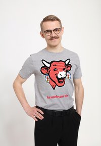 Šedé tričko s grafikou červené krávy s puntíky a naušnicemi, text zní "la vache qui rit", doplněné černými kalhotami.