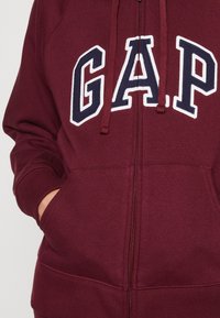 Felpa con zip di color bordeaux con logo "GAP" ricamato bianco e blu navy. Presenta cordini, tasche a marsupio e polsini a costine.