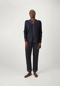 Kvinne står barføtt iført et langermet pysjamassett i marineblått med hvite detaljer på kragen, mansjettene og buksehemmen mot en ensfarget bakgrunn.