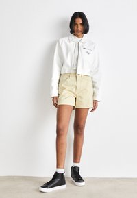 Giacca corta bianca con zip, pantaloni corti beige e sneakers alte nere. Presenta una texture intrecciata e un sottile logo CK sulla giacca.