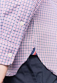 Overhemd met knopen in een rood, wit en blauw ruitpatroon, met een figuuraccentuerend ontwerp en zijsplitten. Witte knopen en contrasterende binnenstof zichtbaar.